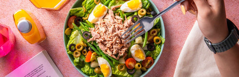 A person holding a fork against a bowl of Salad Nicoise in the middle with Joel's Place powershots and an opened book on the left. 