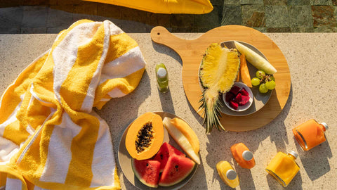 An assortment of summer fruits and Joel's Place powershots on the poolside with a yellow and white striped towel on the side. 