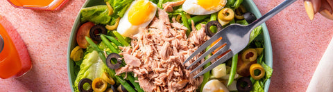 A fork hovering over a Joel's Place salad, next to a glass of orange juice and a power shot bottle, on a pink tabletop