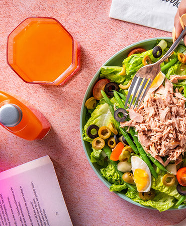 A fork hovering over a Joel's Place salad, next to a glass of orange juice and a power shot bottle, on a pink tabletop