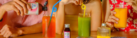 Three people happily drinking colorful, healthy beverages with swirly straws at a table against a verdant background