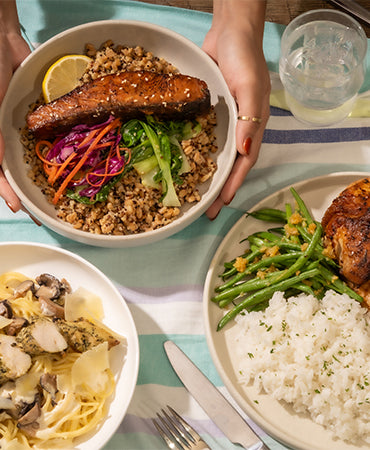 Hands holding a Joel's Place rice bowl meal next to two plates of Joel's Place chicken and pasta dishes, a glass of water, and dip, on top of a striped tablecloth.
