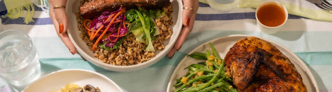 Hands holding a Joel's Place rice bowl meal next to two plates of Joel's Place chicken and pasta dishes, a glass of water, and dip, on top of a striped tablecloth.