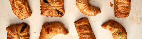 This image displays a flatlay of freshly baked, laminated dough pastries, characterized by their golden, flaky, and buttery layers, that include cronadas, empanadas, and filled pastries,  available at Joel’s Place. 