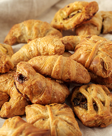 This image displays a variety of freshly baked, laminated dough pastries, characterized by their golden, flaky, and buttery layers, that include cronadas, empanadas, and filled pastries, available at Joel’s Place. 