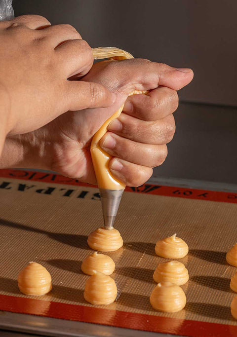 A photo of a hand piping choux pastry dough on a baking mat