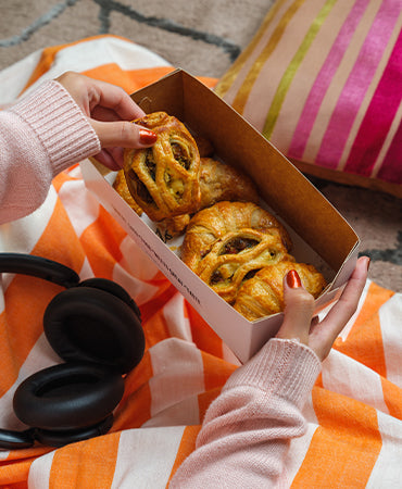A person picking a flaky pastry from a Joel's Place box during a cozy rainy day at home, ideal feel-good food for gloomy days