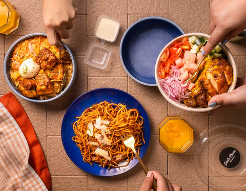 Table set up featuring Joel's Place's Chef Joy's Adobowl on the left, Chicken Shawarma Rice Bowl on the right, and Pasta Bolognese in the middle with three hands holding utensils and getting a serving. 
