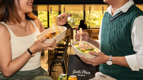 Two people holding Joel's Place ready to eat meals. The girl on the left is holding a container of Shrimp Aglio Olio pasta while the guy on the right side is holding a container of Salad Nicoise. 