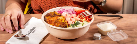 A woman holding a Joel's Place powershot against her chest with a bowl of Joel's Place Chicken Shawarma placed in front with a napkin and spoon on the left, and a garlic sauce on the right. 