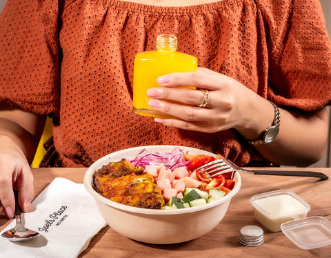 A woman holding a Joel's Place powershot against her chest with a bowl of Joel's Place Chicken Shawarma placed in front with a napkin and spoon on the left, and a garlic sauce on the right. 