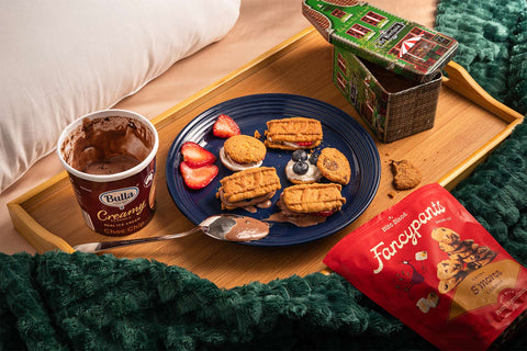 A wooden tray placed on top of a bed with a white pillow and a green throw. On top of the tray is a blue plate filled with cookie sandwiches with whipped cream, ice cream, and fruit. There is an open tub of Bulla Choc Chip ice cream, a green can of Vermeiren Traditional Biscuits, and a bag of Fancypants Crispy S'mores Cookies.