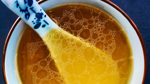 A close up photo of a bowl of golden broth with a ceramic spoon, showing oil droplets on the surface