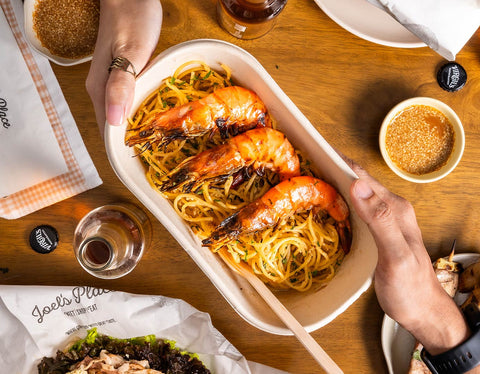 Two hands holding a container filled with Joel's Place Shrimp Aglio Olio. It is against a wooden table with sandwiches, sauces, drinks, and napkins on the side. 