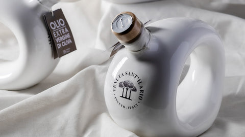 A white, uniquely shaped bottle of Tenuta Sant’ilario Pineto extra virgin olive oil on top of a wrinkled white fabric, near another bottle of the same brand and kind of olive oil.